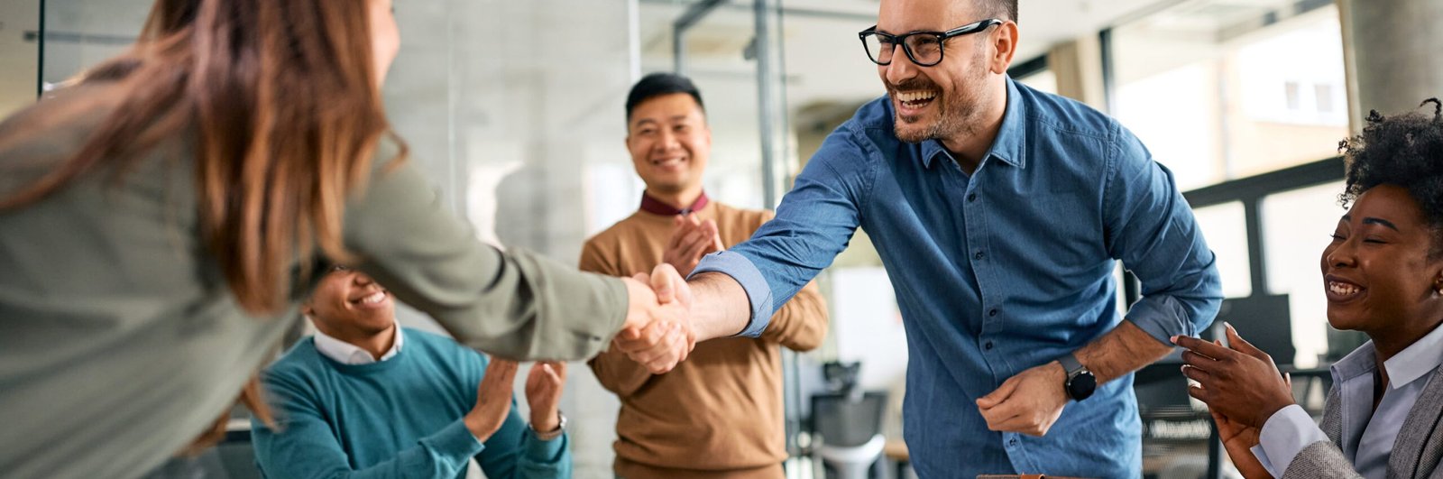 Happy businessman handshaking with female colleague during a meeting in the office while others are applauding.